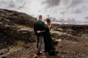 Ein Paar steht umarmt auf felsigem Gelände unter einem bewölkten Himmel. Die Frau trägt ein fließendes schwarzes Kleid und der Mann einen dunklen Anzug. Sie blicken abgewandt auf die zerklüftete Landschaft.