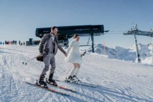 Ein Paar fährt Händchen haltend einen verschneiten Hang hinunter. Der Mann trägt einen braunen Anzug und die Frau ein weißes Kleid. Hinter ihnen ist ein Skilift und ein klarer blauer Himmel mit schneebedeckten Bergen im Hintergrund.