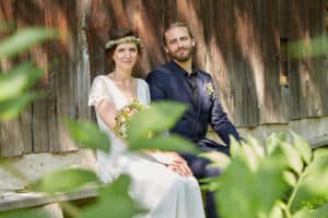 Braut und Bräutigam sitzen auf einer Bank, umgeben von üppigem Grün. Die Braut, strahlend in ihrem be.lichteten weißen Kleid und ihrer Blumenkrone, hält einen Blumenstrauß. Der Bräutigam ergänzt sie in einem dunklen Anzug. Zusammen verleihen sie der rustikalen Holzkulisse Eleganz und schaffen eine bezaubernde und natürliche Kulisse.