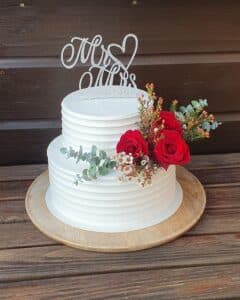 Eine zweistöckige Torte mit weißem Zuckerguss auf einem Holzbrett, geschmückt mit roten Rosen, kleinen Blumen und Grünpflanzen. Oben ist ein silberner „Mr & Mrs“-Aufsatz mit einem Herz platziert, vor einem dunklen Holzhintergrund.