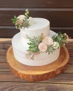 Eine zweistöckige, halbnackte Torte auf einer Holzplatte, dekoriert mit blassrosa und weißen Rosen, Grünpflanzen und kleinen rosa Blumen vor einem rustikalen Holzhintergrund.