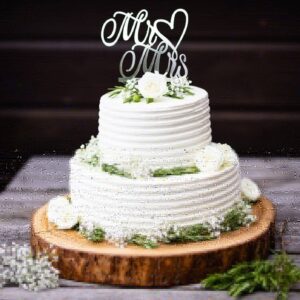 Die Cake Deluxe ist eine zweistöckige weiße Hochzeitstorte mit gerippter Glasur und Blumendekoration. Der „Mr & Mrs“-Aufsatz ist mit einem Herz verziert. Er steht auf einem Holzsockel, umgeben von Grünpflanzen und weißen Blumen, vor einem dunklen Holzhintergrund.