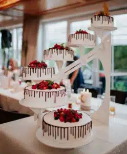 Auf einem mehrstöckigen Tortenständer stehen fünf weiße Torten, die mit Schokoladentropfen dekoriert und mit verschiedenen Beeren garniert sind. Der Ständer steht auf einem weiß gedeckten Tisch in einem hellen Raum mit Fenstern und unscharfem Hintergrund.