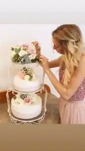 Eine Frau mit langen blonden Haaren verziert eine zweistöckige Torte mit rosa und weißen Rosen. Die Torte steht auf einem verzierten weißen Ständer. Sie trägt ein glitzerndes rosa Oberteil und einen Tüllrock. Der Hintergrund ist eine schlichte weiße Wand.