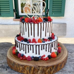 Ein zweistöckiger Kuchen auf einer Holzplatte, mit frischen Beeren belegt und mit Schokoladentropfen überzogen. Ein dekorativer Aufsatz besteht aus ineinander verschlungenen Herzen mit der Aufschrift „Mr & Mrs“. Der Hintergrund zeigt eine strukturierte Wand und grüne Fensterläden.